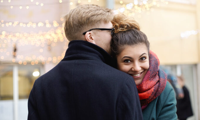 Sweet moment of true love. Romantic young couple in winter clothes cuddling outdoors: Hispanic girl with nose ring and blonde Caucasian guy in eyeglasses hugging on street decorated with light garland